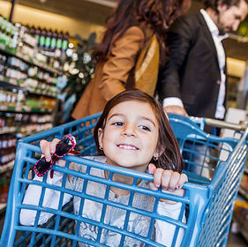 A child sits in a shopping cart as her parents in the background look at the groceries on the aisle.