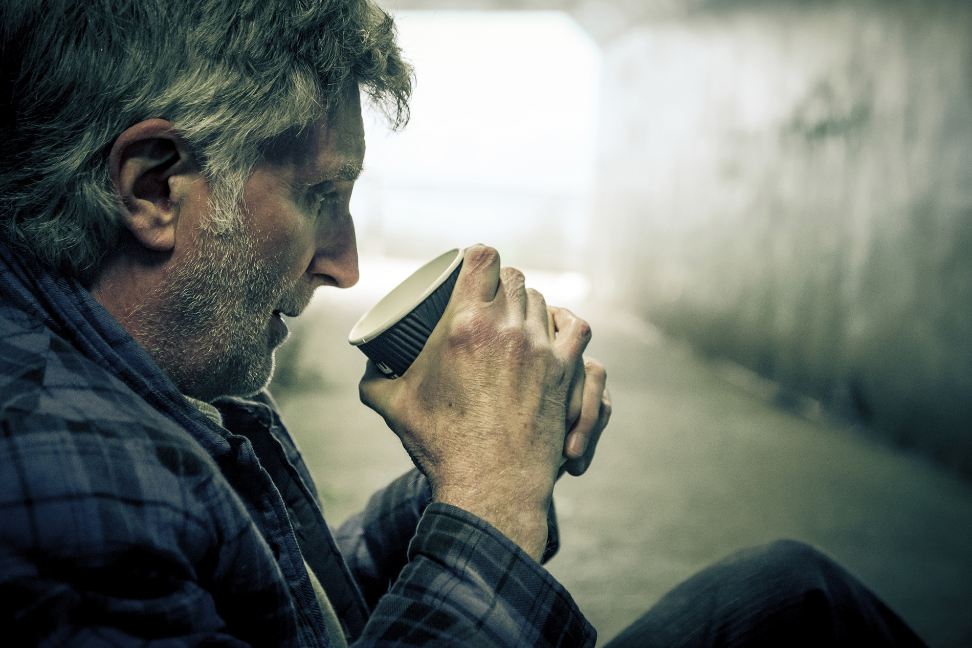 An older man is looking into a coffee cup. He's sitting in a dark place, under a bridge.