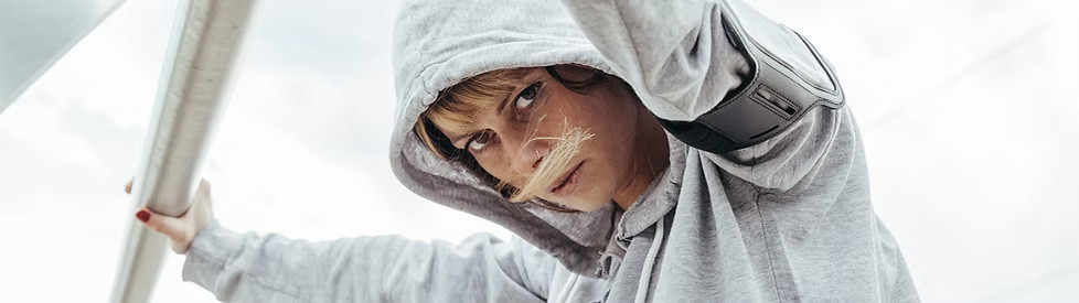 a girl in a light gray hoodie stops her jog to stretch her arms, holding a pole above her.