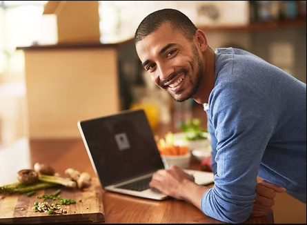 A man is in the kitchen leaning over his laptop and smiling back towards the camera.